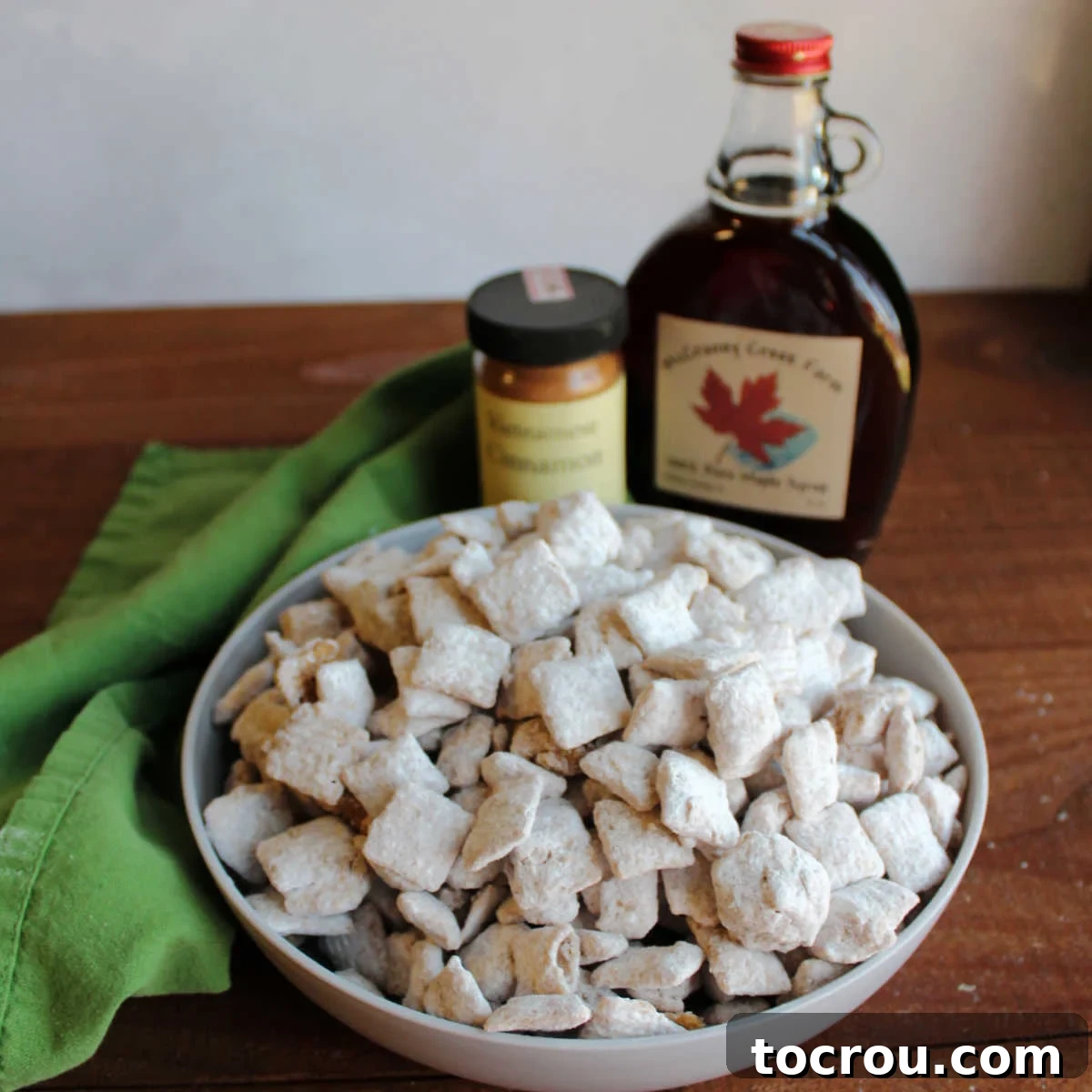 Bowl of powdered sugar coated French Toast Puppy Chow in front of bottle of maple syrup and cinnamon.