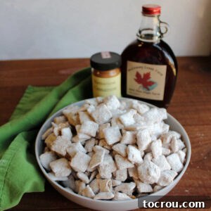Bowl of powdered sugar coated French Toast Puppy Chow in front of bottle of maple syrup and cinnamon.