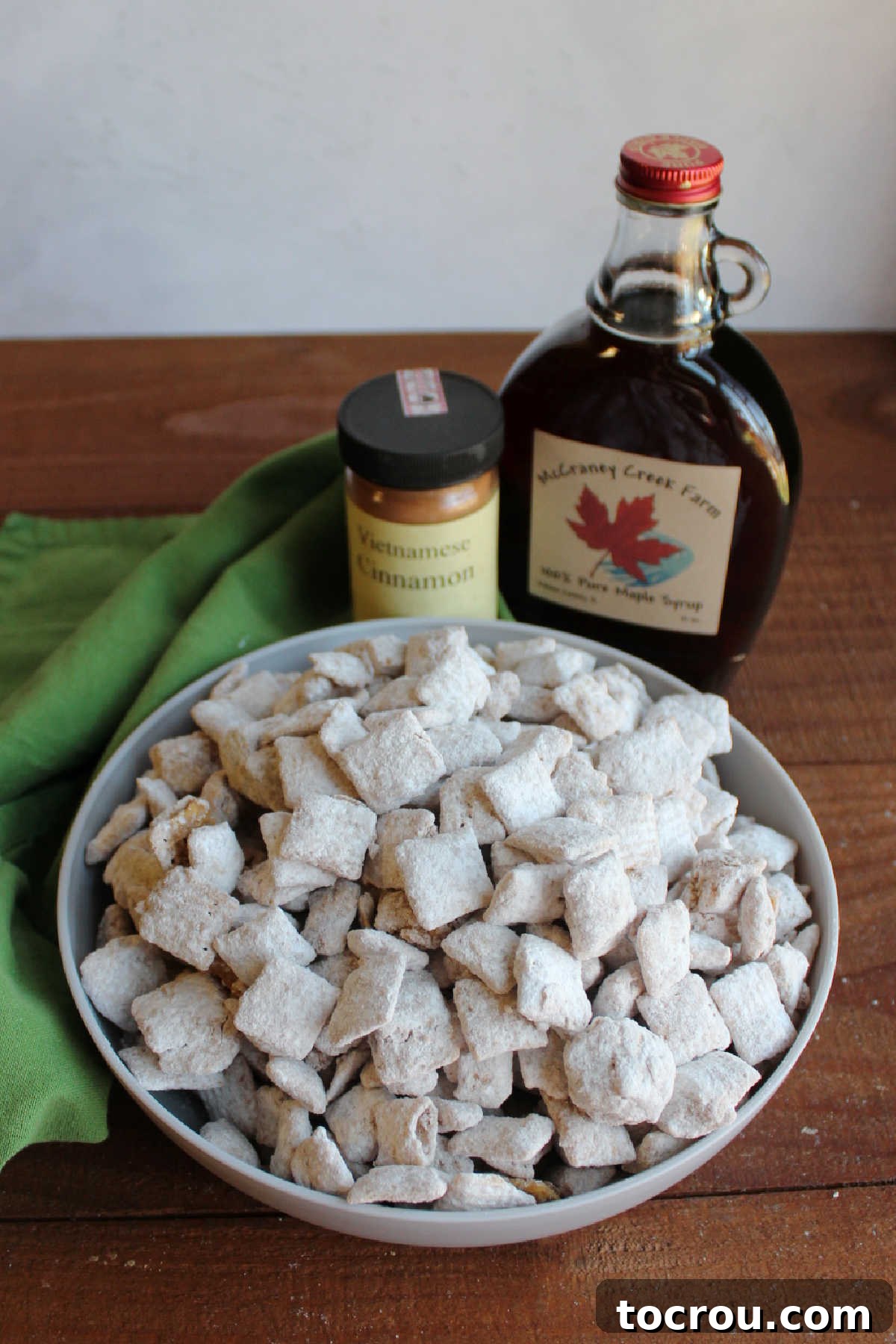 Bowl of maple cinnamon puppy chow next to bottle of pure maple syrup and jar of cinnamon.