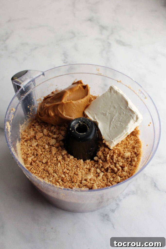 Cookie crumbs, softened cream cheese, and creamy peanut butter in a food processor, ready to be blended.