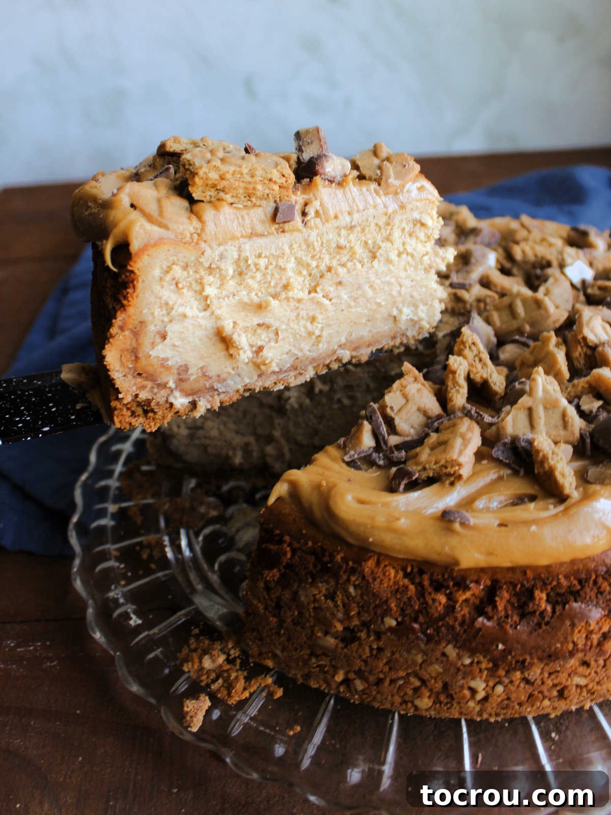 A Slice Ready to Serve Lifting piece of peanut butter cheesecake off the serving platter showing creamy filling, soft peanut butter topping, and bits of nutter butters and heath bars on top.