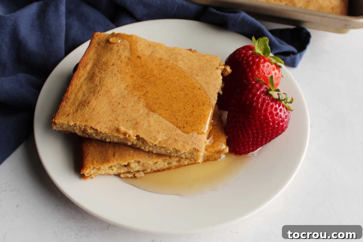 Two pieces of golden-brown whole wheat banana sheet pan pancakes stacked on a plate, generously drizzled with maple syrup. Fresh strawberries are placed beside the pancakes, highlighting a delicious and easy breakfast option.