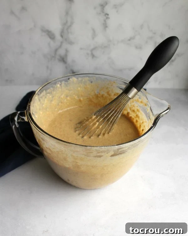 Clear glass batter bowl filled with a smooth, golden whole wheat banana pancake batter, accompanied by a whisk ready for mixing. This image suggests the simple preparation process.
