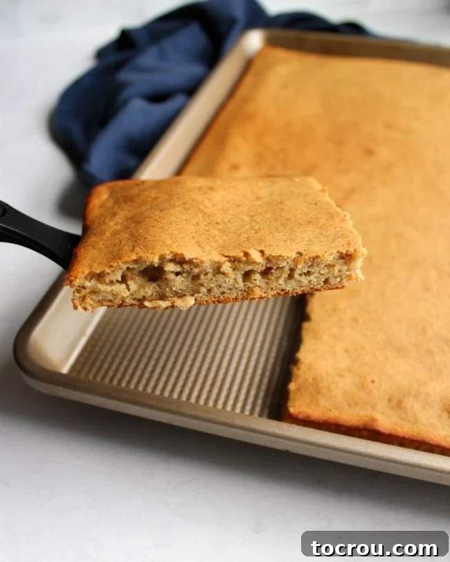 A spatula gently lifts a square piece of fluffy banana sheet pan pancake, revealing its soft, airy texture inside. The golden-brown surface indicates perfect baking.