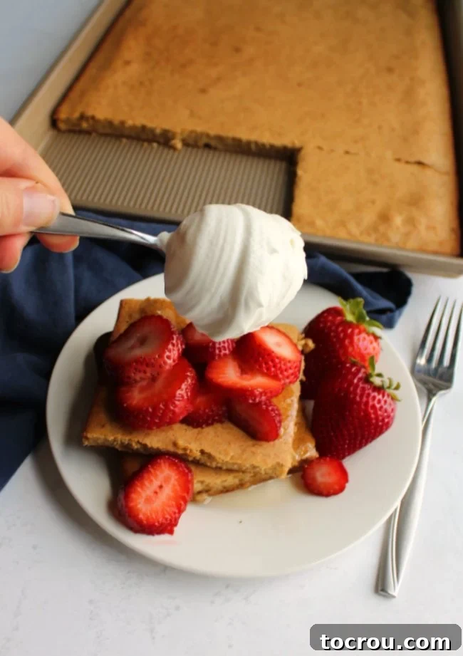 A plate of golden banana sheet pan pancake squares, beautifully topped with vibrant fresh strawberry slices and a generous dollop of creamy whipped cream. The presentation elevates a simple breakfast into a delightful treat.