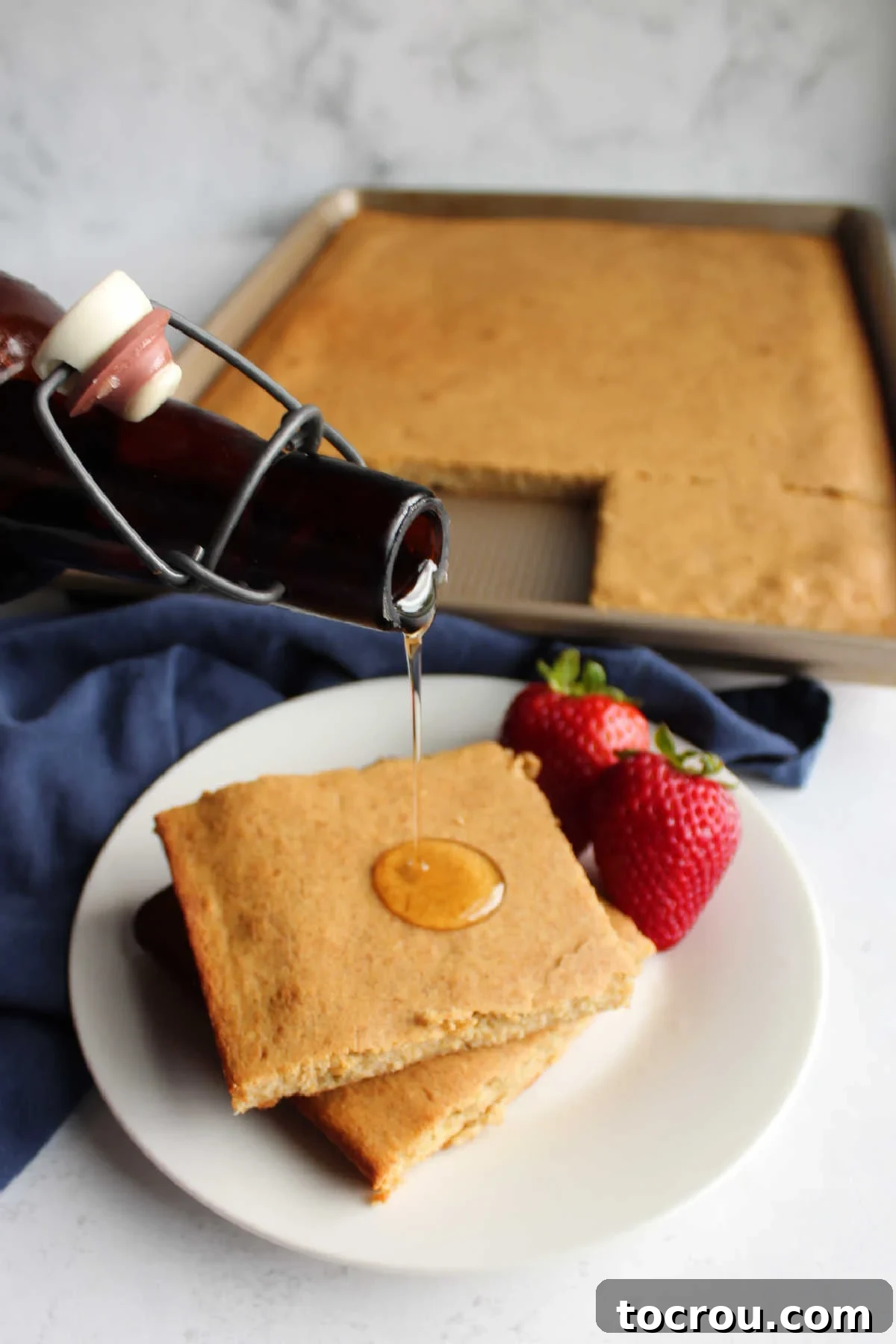 A delightful stack of square banana sheet pan pancakes being generously drizzled with warm, golden maple syrup, creating an enticing visual of a sweet and satisfying breakfast.
