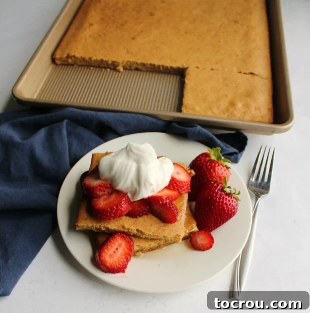 A stack of square sheet pan pancakes artfully topped with fresh strawberry slices and a generous dollop of maple whipped cream, with the rest of the baked sheet pan pancake visible in the background, showcasing its golden perfection.