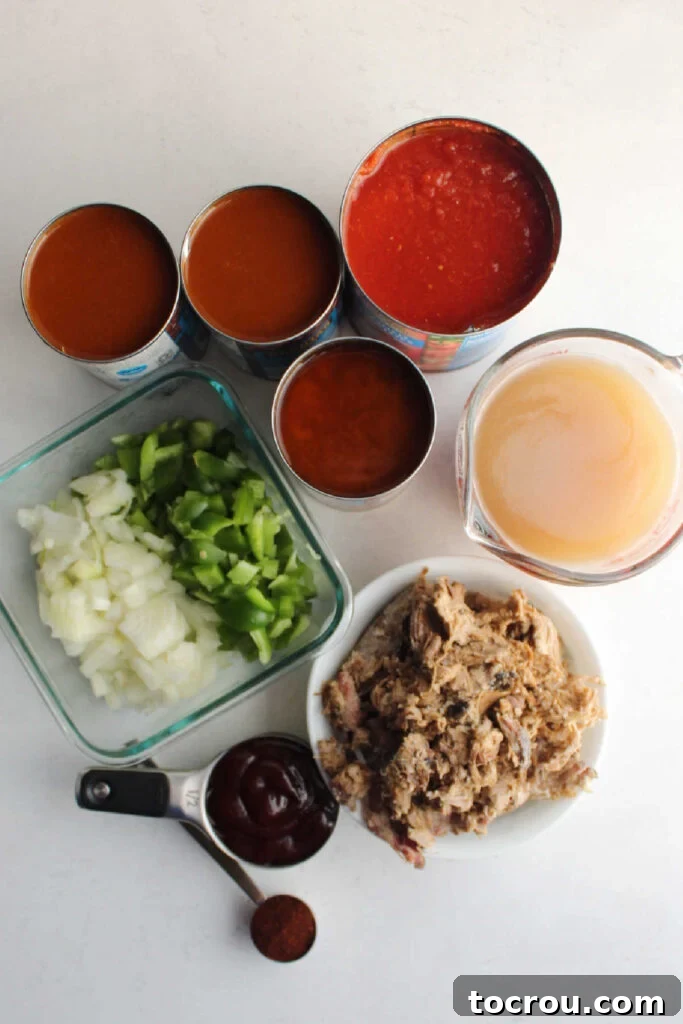 Assorted ingredients laid out for slow cooker pulled pork chili, including cans of beans and tomatoes, a bowl of diced onions and peppers, a measuring cup of beef broth, and a portion of cooked pulled pork, ready for assembly.