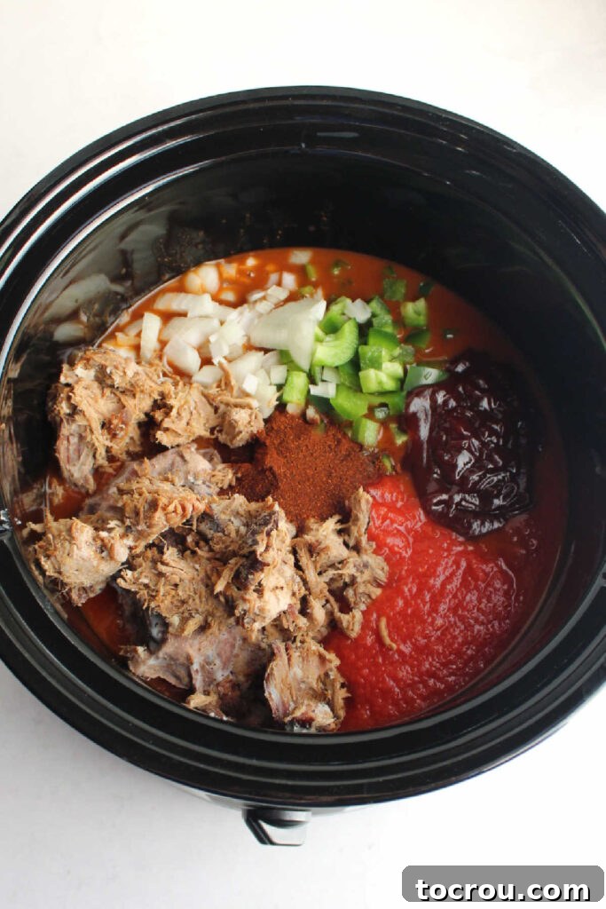 The inside of a crock pot filled with all the uncooked ingredients for pulled pork chili: tender pulled pork, colorful diced bell peppers and onions, various canned beans, and crushed tomatoes, all ready to be simmered.