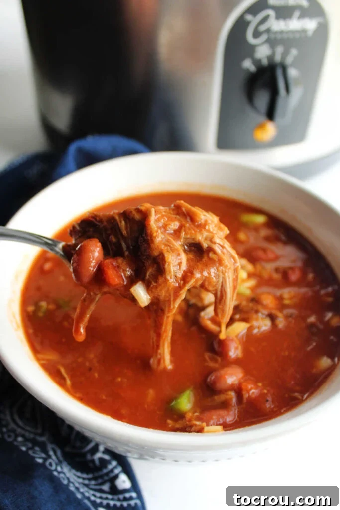 A spoon full of rich, slow-cooked pulled pork chili, showcasing tender meat, beans, and melted cheese, positioned in front of a bubbling crock pot, indicating readiness to serve.