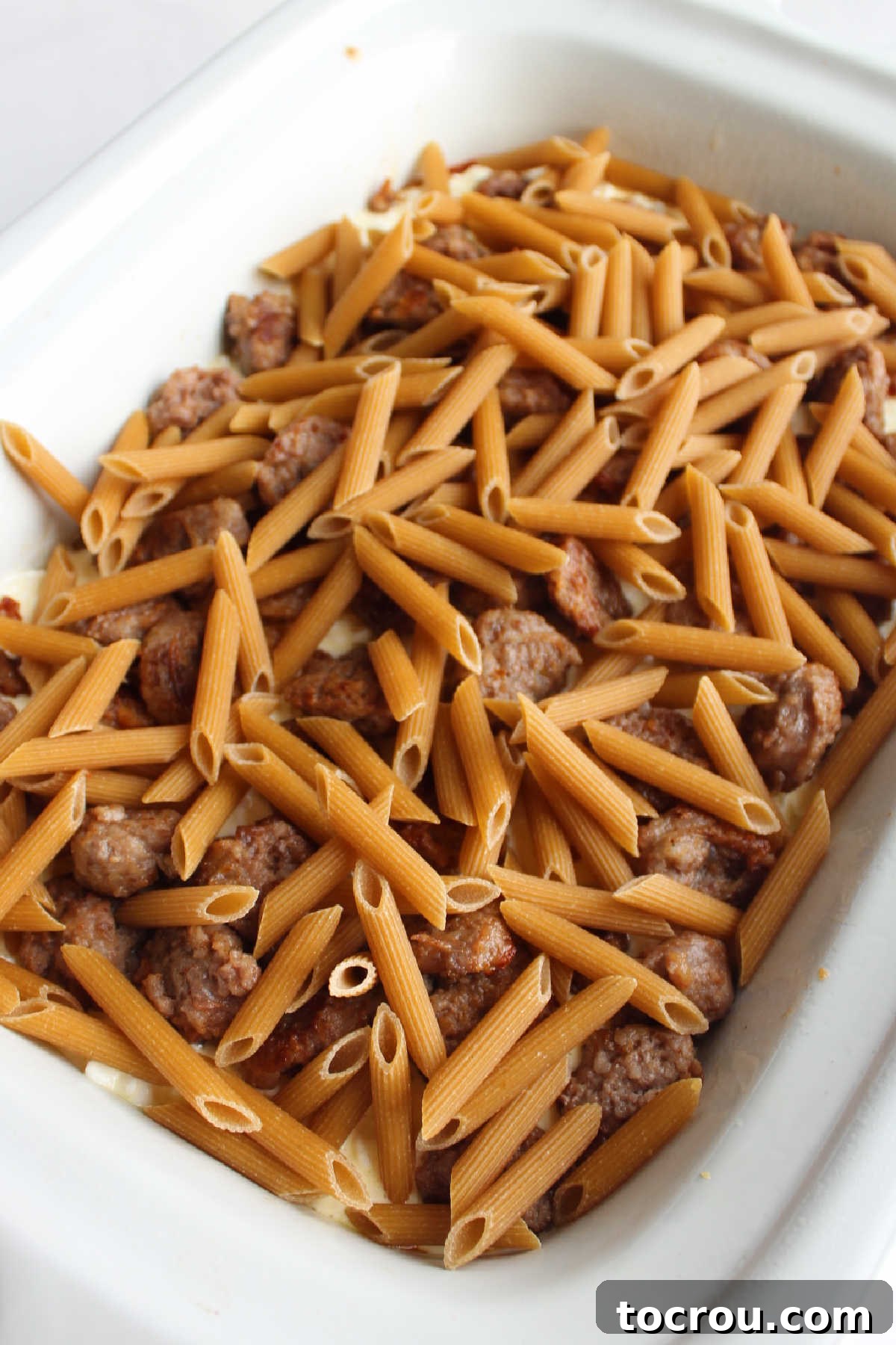 Raw penne pasta layered over cheese and sausage in slow cooker.