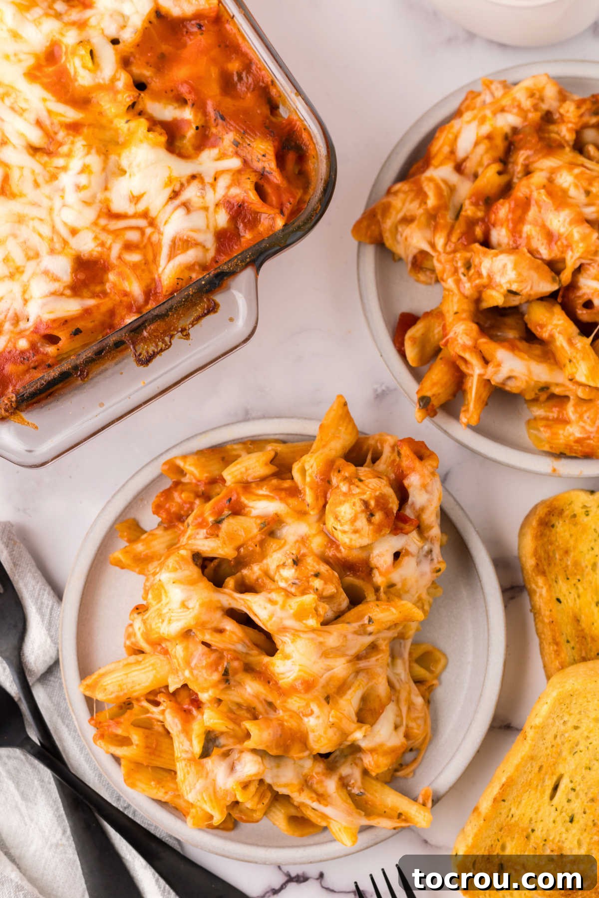 Dump-and-Bake Chicken Pasta Casserole 7 Two dinner plates with baked chicken and pasta casserole next to baking dish and some slices of garlic bread.