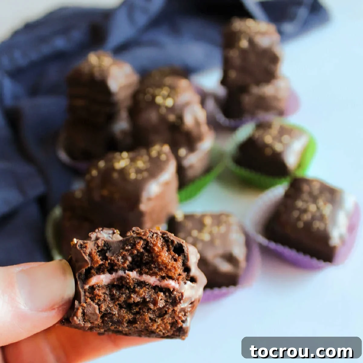 A hand holding a perfectly crafted chocolate petit four, revealing its moist chocolate cake interior layered with vibrant pink strawberry buttercream, all encased in a smooth dark chocolate shell.