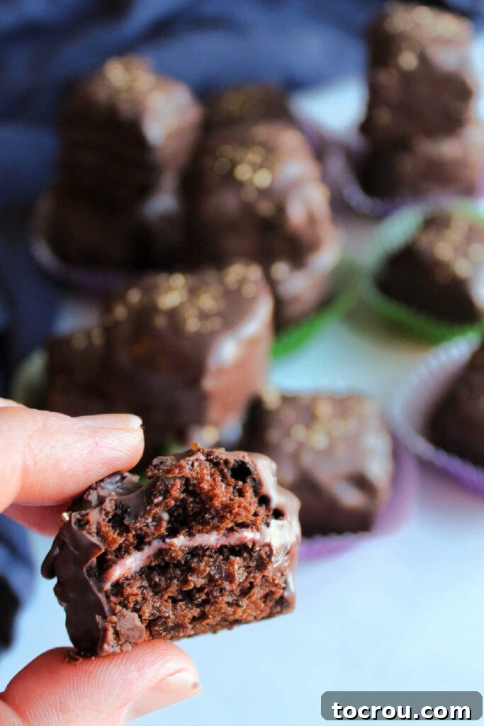 A close-up view of a chocolate petit four sliced open, revealing its distinct layers: a rich chocolate cake base, a creamy pink strawberry buttercream filling, and a perfect coating of dark chocolate.