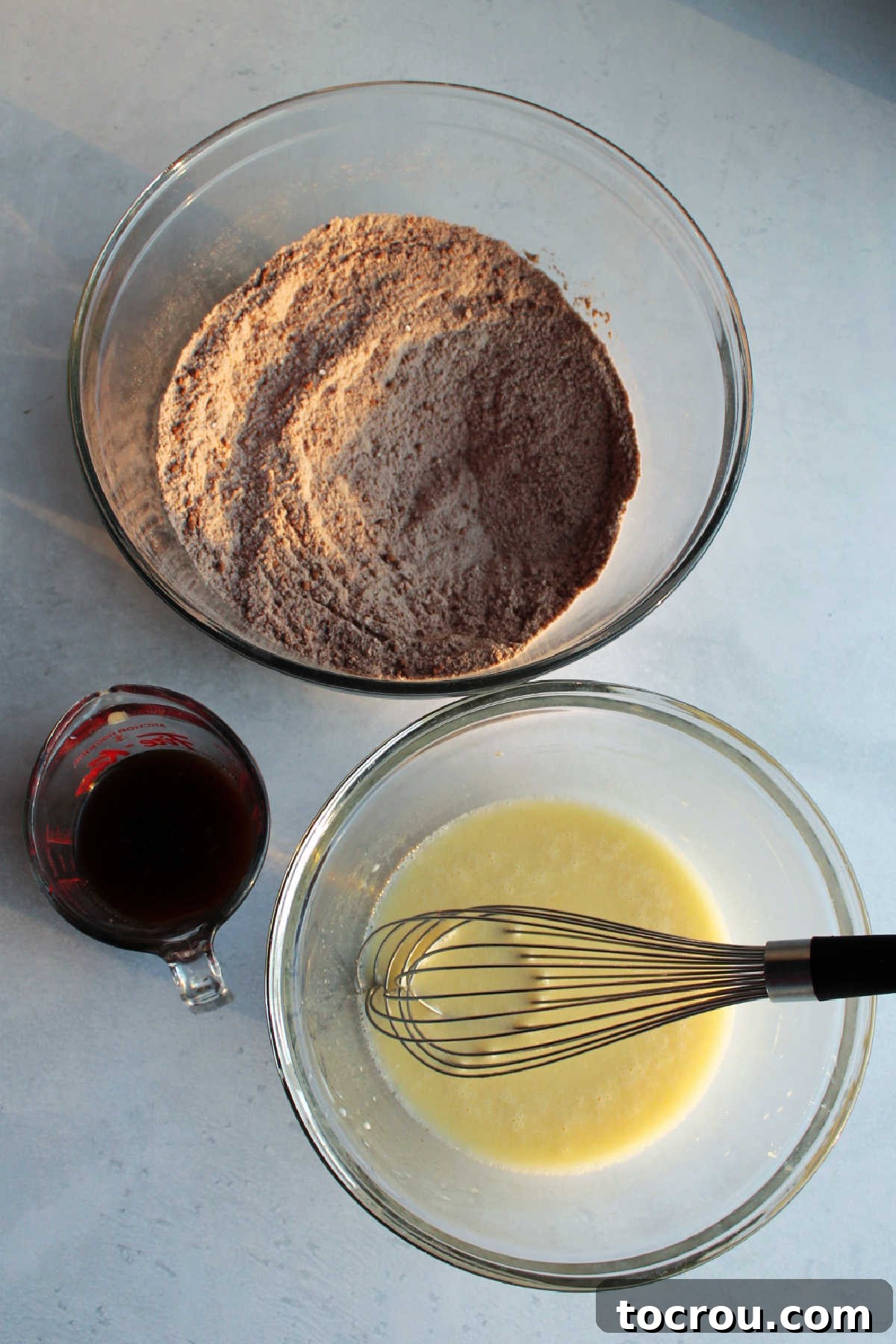 A collection of essential ingredients for chocolate cake batter, including a bowl of dry ingredients, a bowl of wet ingredients, and a measuring cup of hot coffee, all ready for mixing.