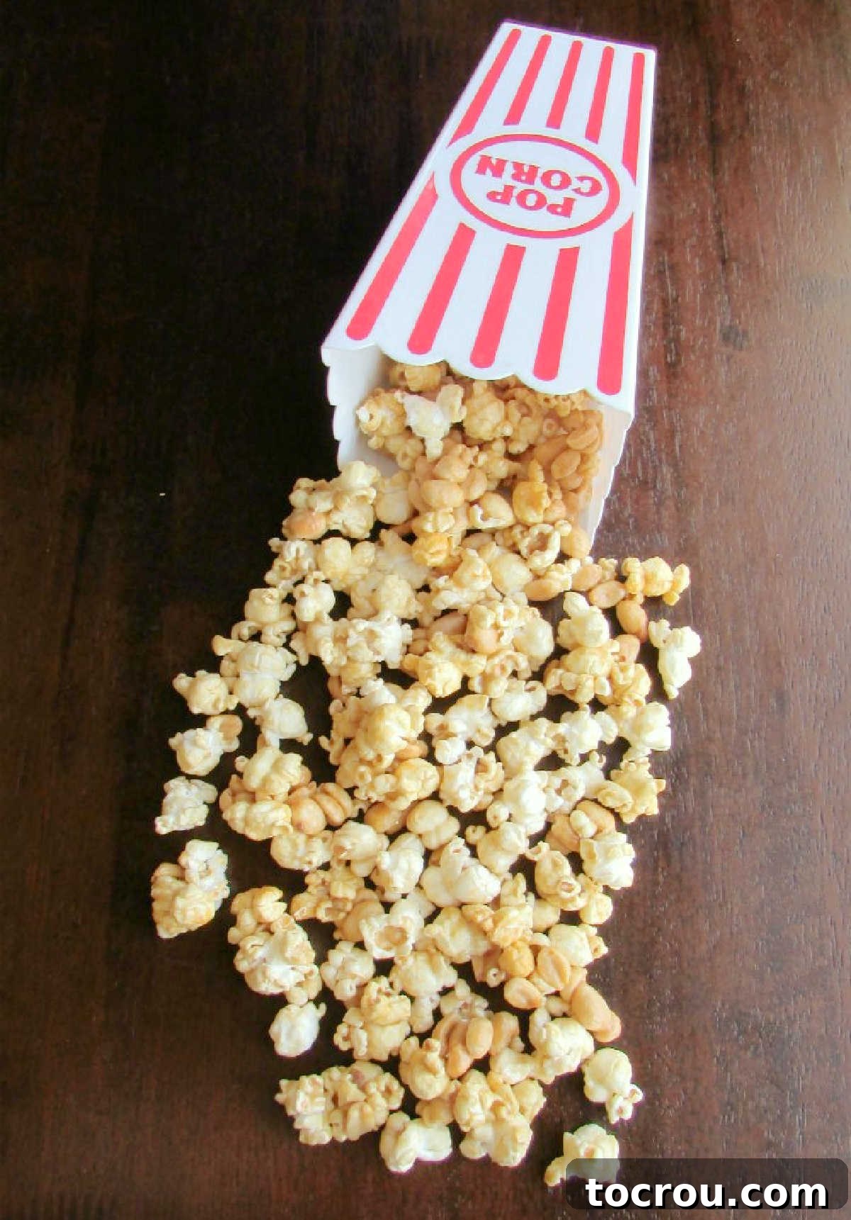 A red and white striped popcorn container with caramel corn spilled out, clearly showing individual pieces coated in a shiny, crunchy caramel layer with peanuts interspersed.