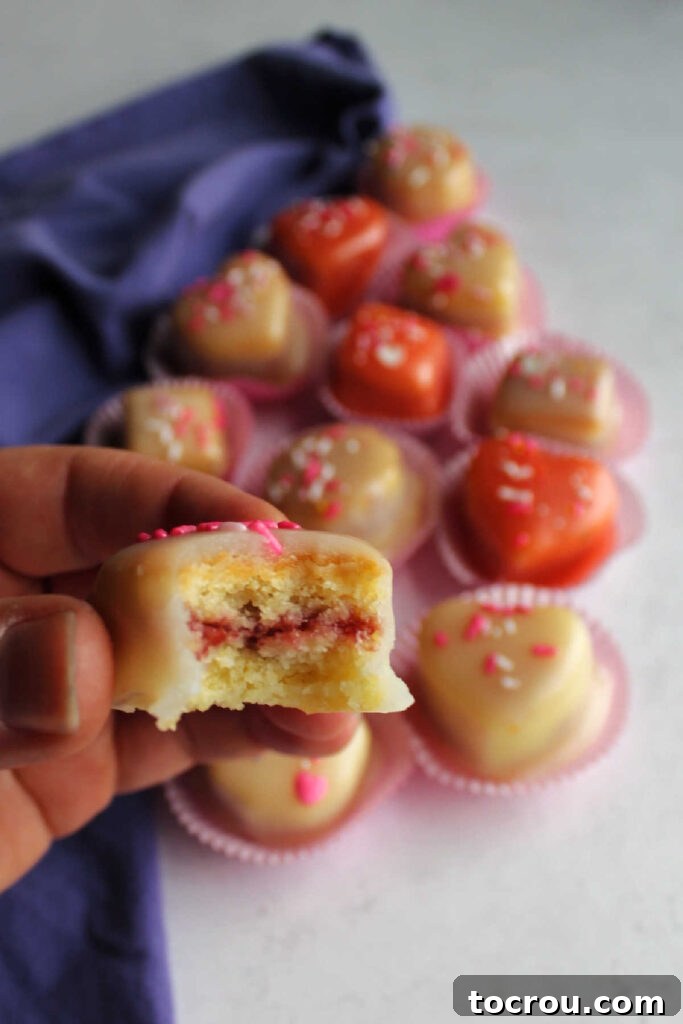 Hand holding a petit four with a bite taken out, showing the raspberry center.