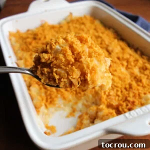 A serving spoon scoops a generous portion of French onion cheesy potato casserole, adorned with a golden brown cornflake crumb topping, promising rich flavor and satisfying texture.
