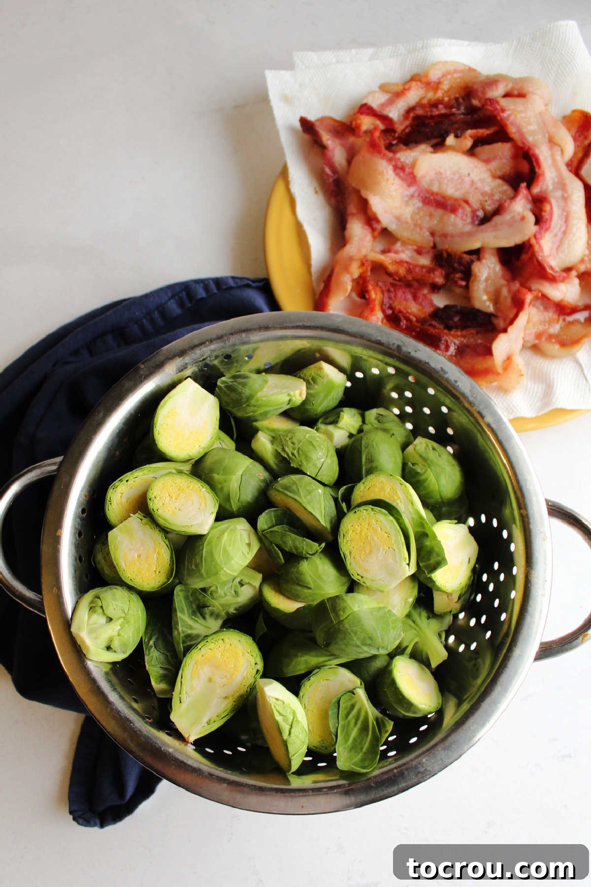 Strainer filled with raw Brussels sprout halves next to a plate of cooked bacon draining on paper towels.