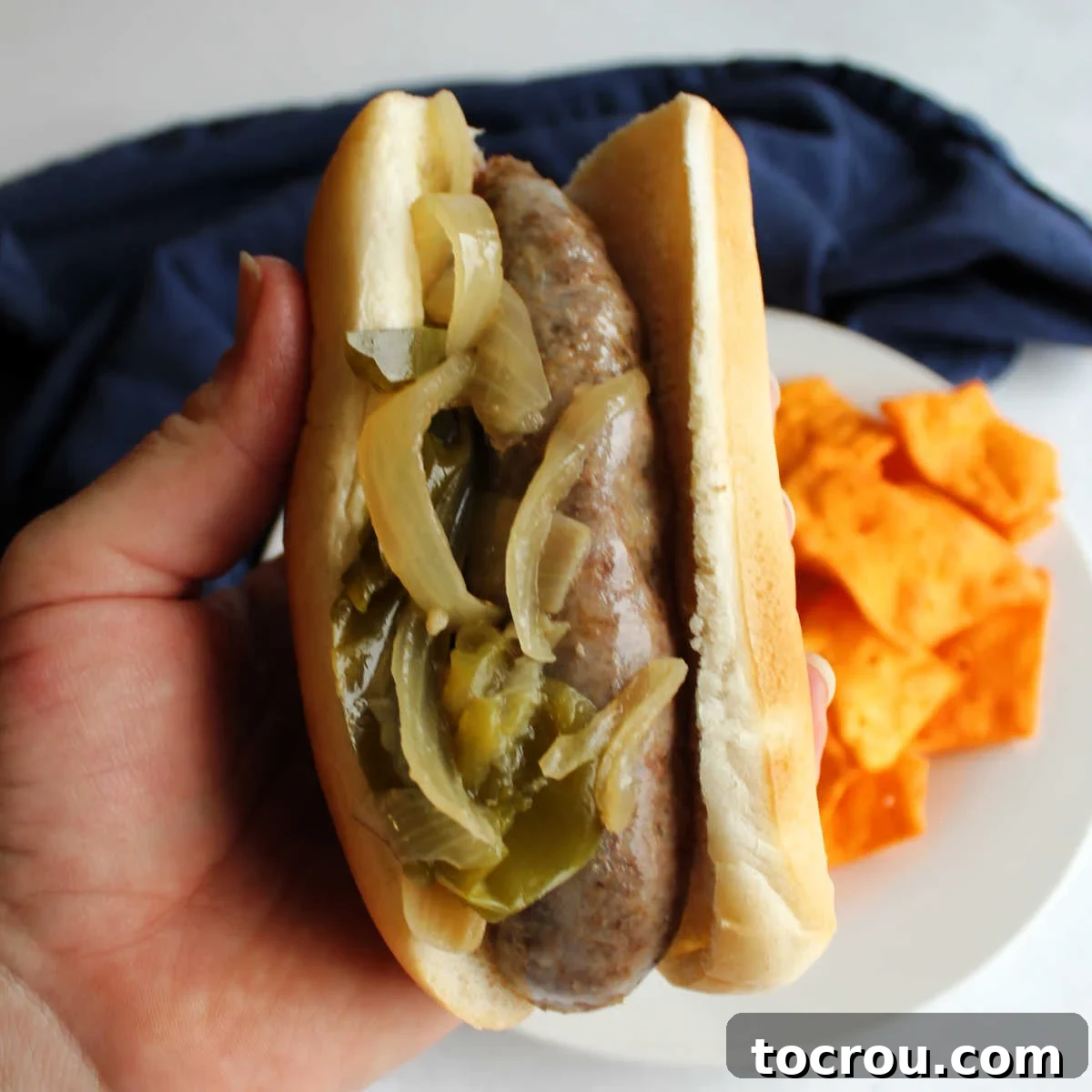 A perfectly cooked bratwurst nestled in a soft bun, generously topped with savory, slow-cooked onions and green bell peppers, held by a hand.