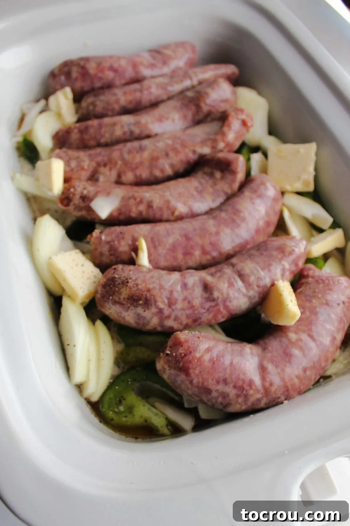 Raw brats, sliced onions, and green bell peppers are layered in a slow cooker with butter, ready for cooking.