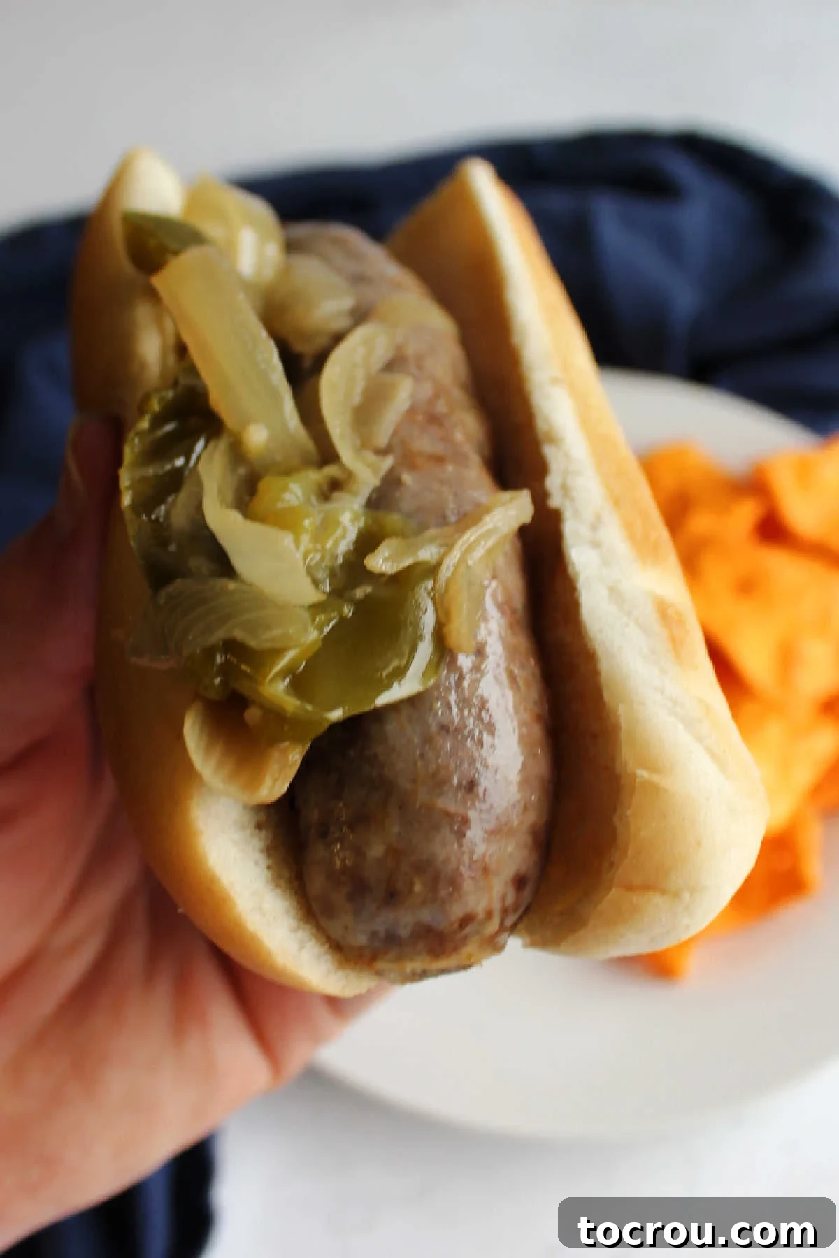 A close-up side view of a hand holding a loaded bratwurst bun, showcasing the juicy brat, onions, and peppers, ready for a delicious bite.