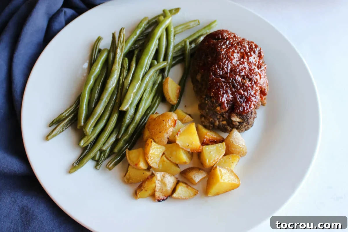 A dinner plate featuring a mini meatloaf topped with a rich sauce, alongside roasted green beans and golden-brown roasted potatoes, with the full sheet pan in the background.