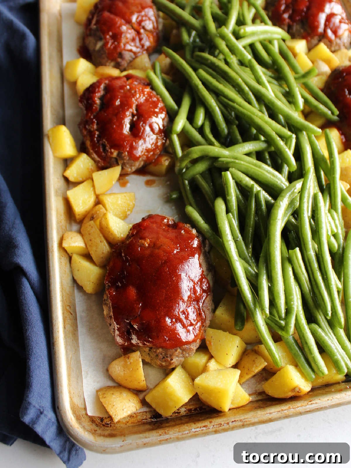 A sheet pan featuring partially cooked mini meatloaves, beautifully coated in a rich, reddish sauce, surrounded by golden cubed potatoes and vibrant fresh green beans.
