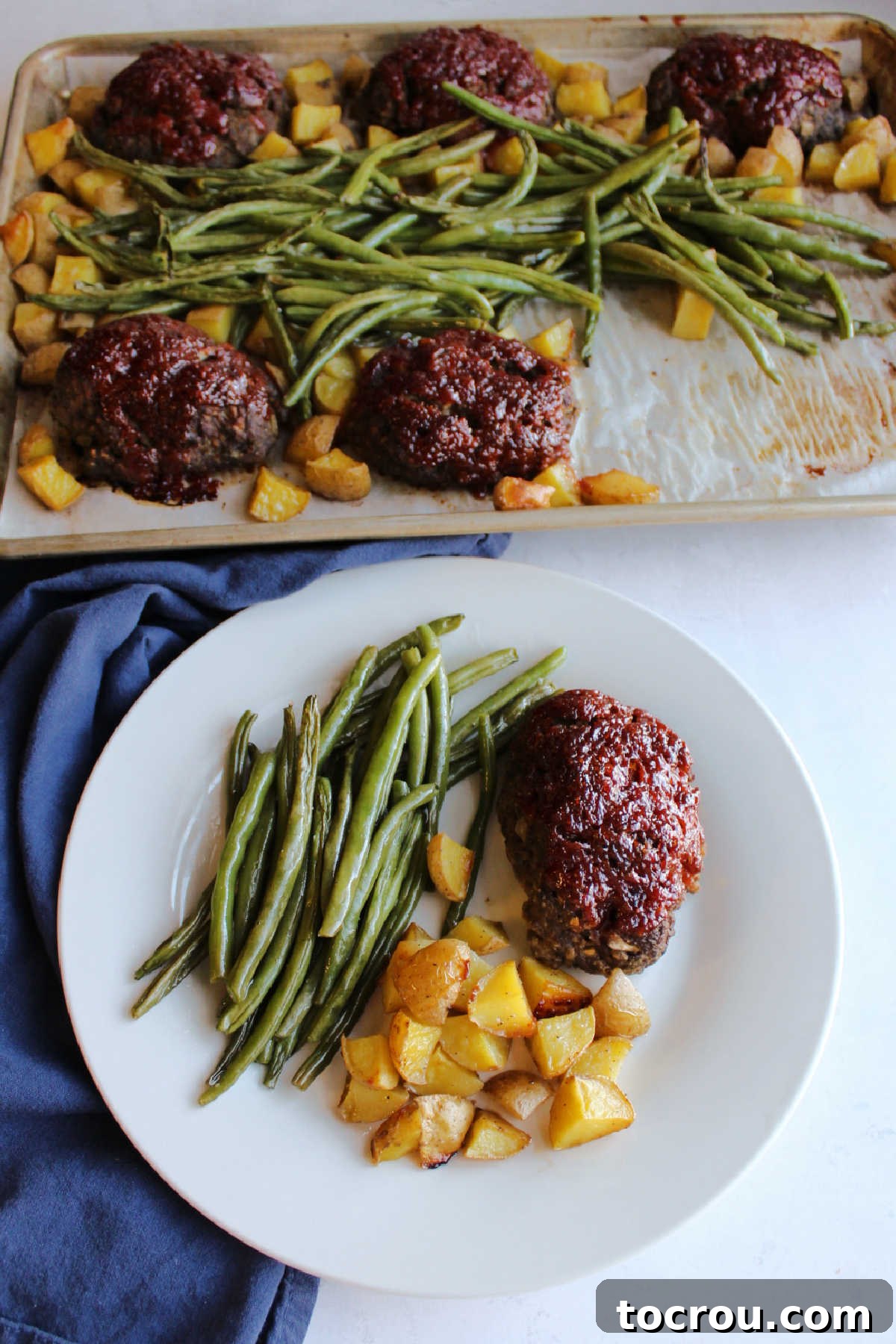 A close-up of a mini meatloaf with glossy red sauce on a dinner plate, accompanied by perfectly roasted potato cubes and fresh green beans, with the remaining sheet pan meal blurred in the background.