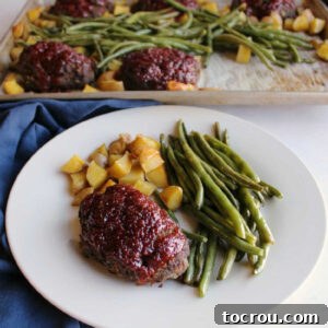 A dinner plate abundantly loaded with a mini meatloaf, crisp green beans, and golden potatoes, with the entire sheet pan visible in the background.