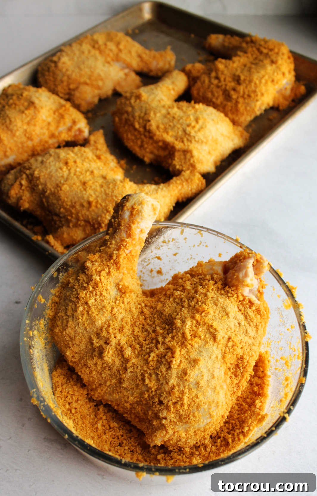 Coating a chicken leg quarter in seasoned cornflake crumbs with tray of chicken ready to bake in the background.