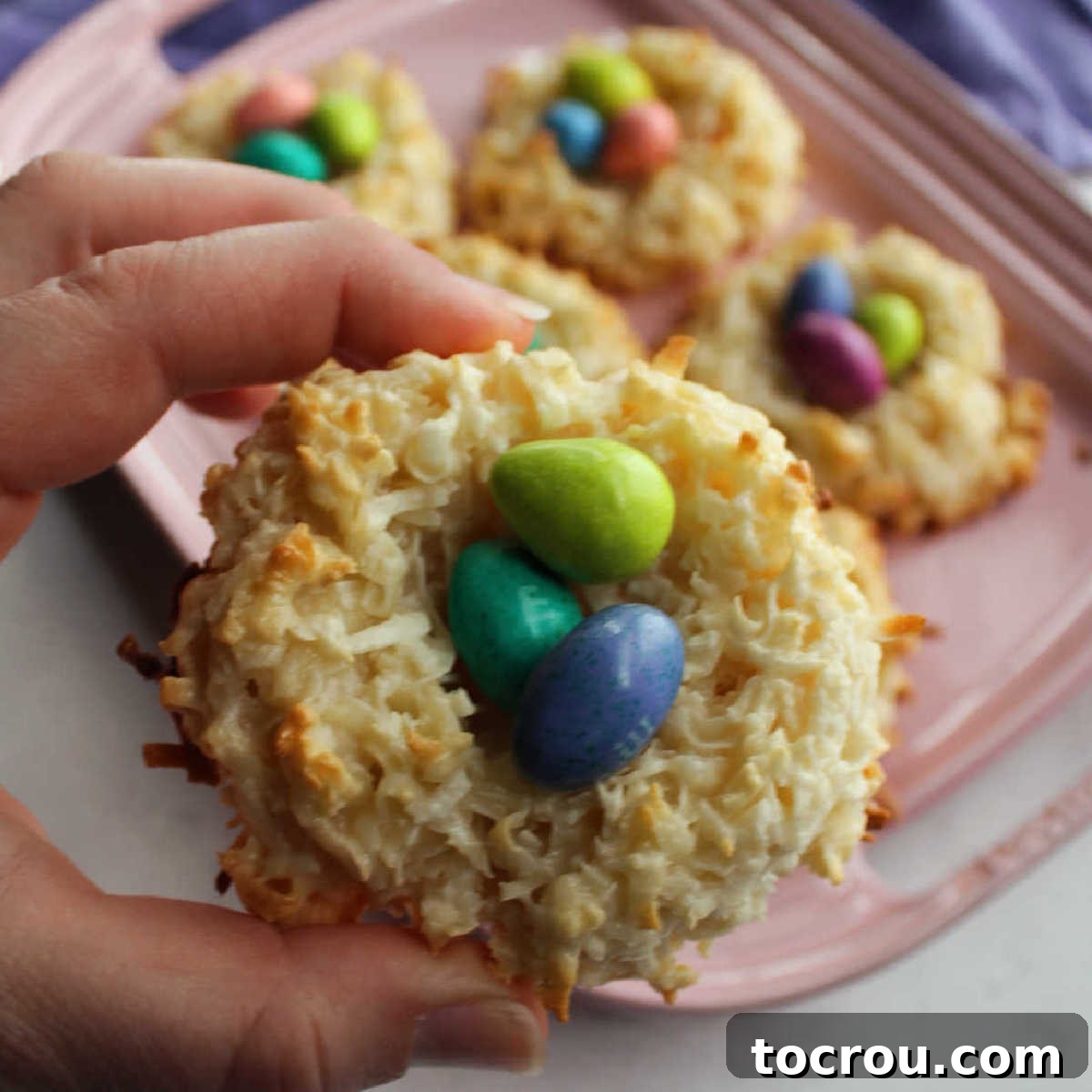 Hand holding a perfectly crafted coconut macaroon nest filled with colorful candy eggs, against a soft-focus background.