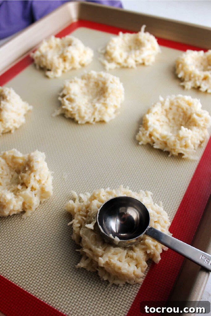 A hand gently pressing the back of a spoon into a mound of coconut macaroon dough on a baking sheet, creating the perfect nest shape before baking.