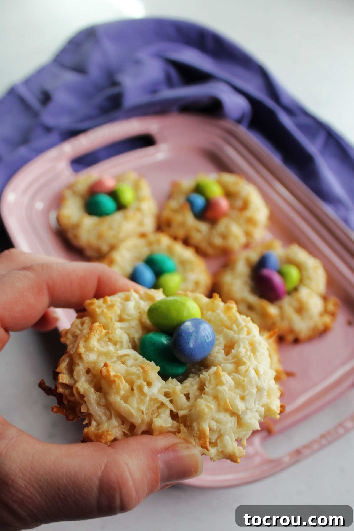 A hand presenting a golden-brown coconut macaroon nest, filled with miniature chocolate eggs, showcasing the delicate balance of crispy coconut and sweet filling.