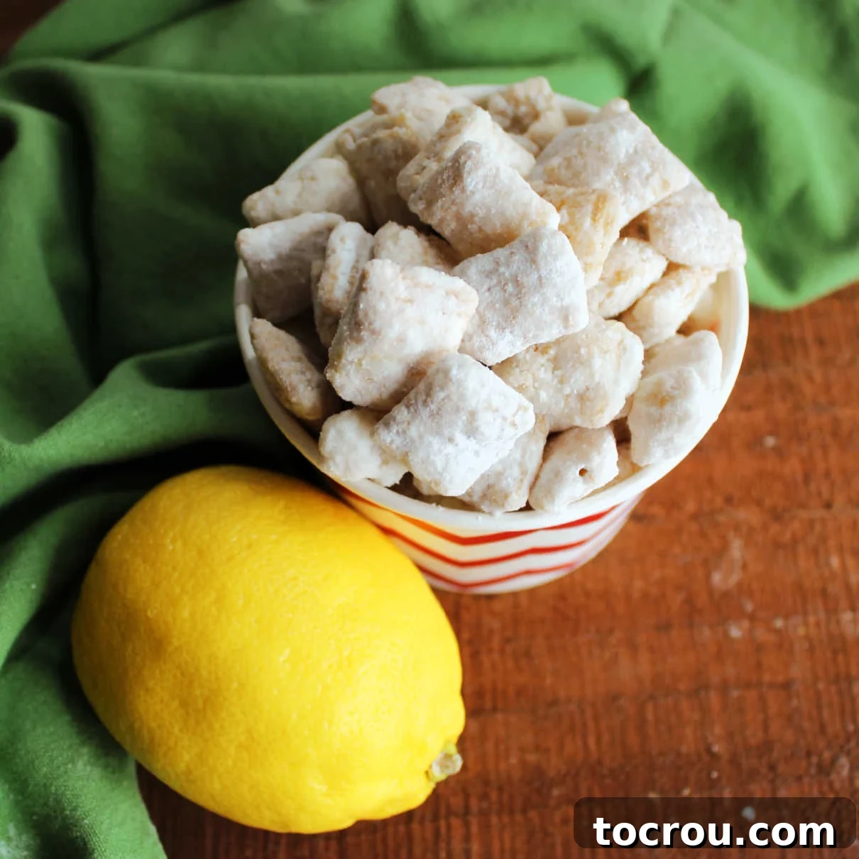 A small bowl of vibrant yellow lemon puppy chow next to a fresh lemon slice, highlighting its refreshing citrus appeal.