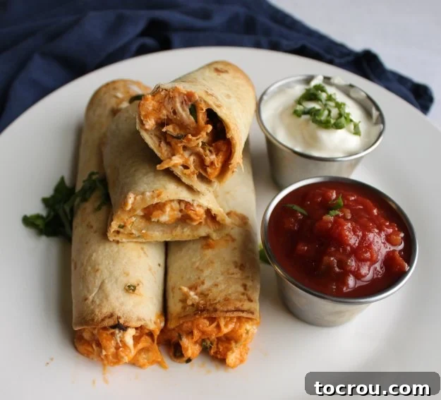 Easy Baked Creamy Chicken Taquitos Recipe Close-up view of a creamy chicken taquito, cut open to reveal the filling, with bowls of sour cream and salsa for dipping in the background.