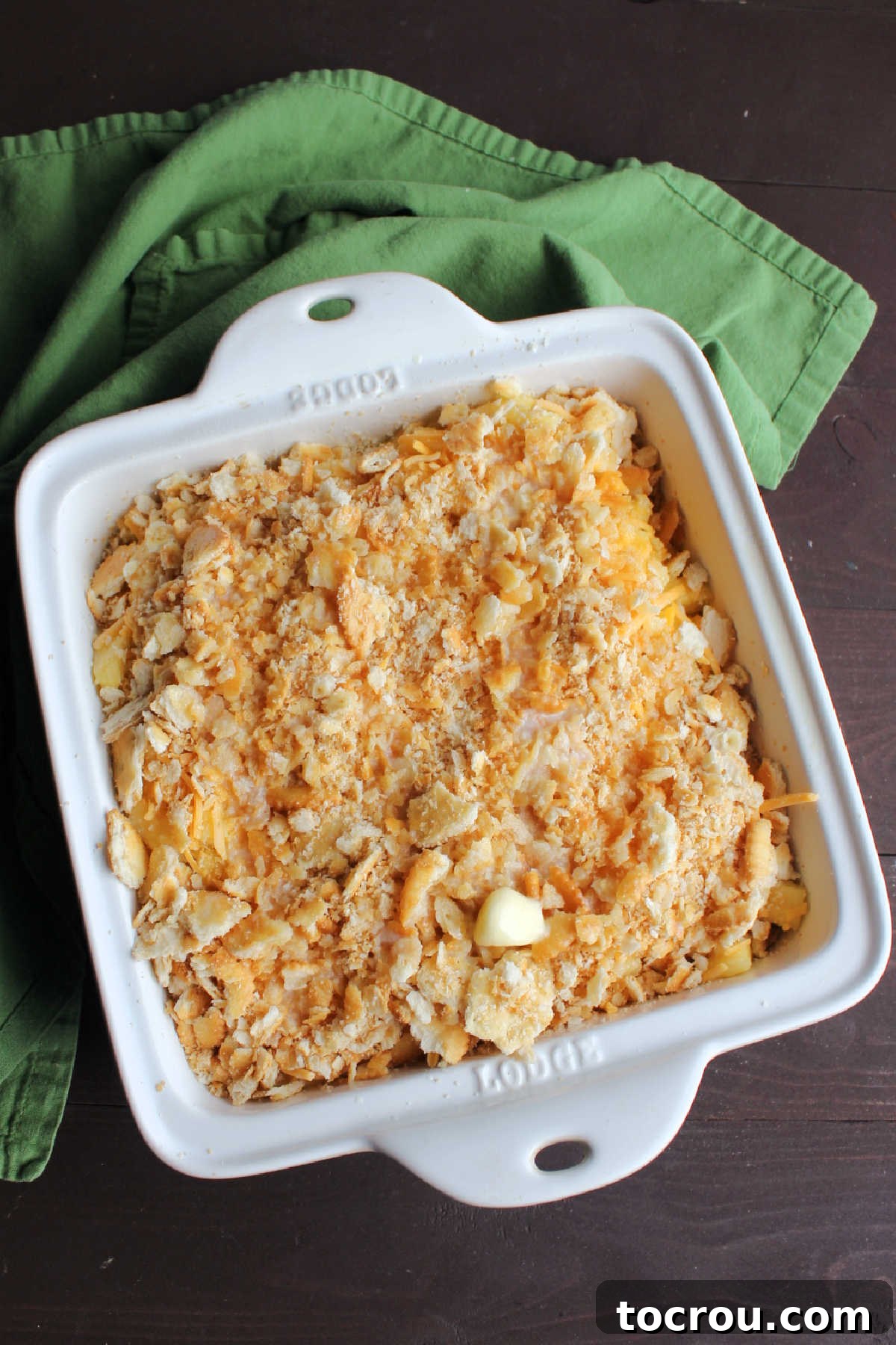 Square baking dish filled with pineapple mixture topped with cracker crumbs and cheddar cheese ready to go in the oven.