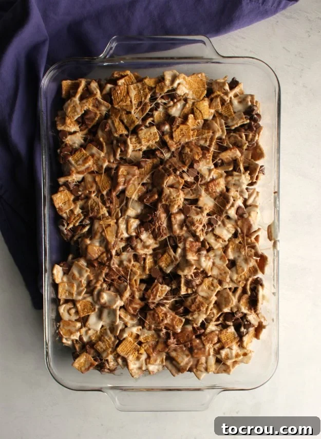 Batch of No-Bake S'mores Bars Glass 9x13-inch baking pan filled with golden s'mores treats, featuring melted marshmallow binding golden grahams cereal and chocolate chips.