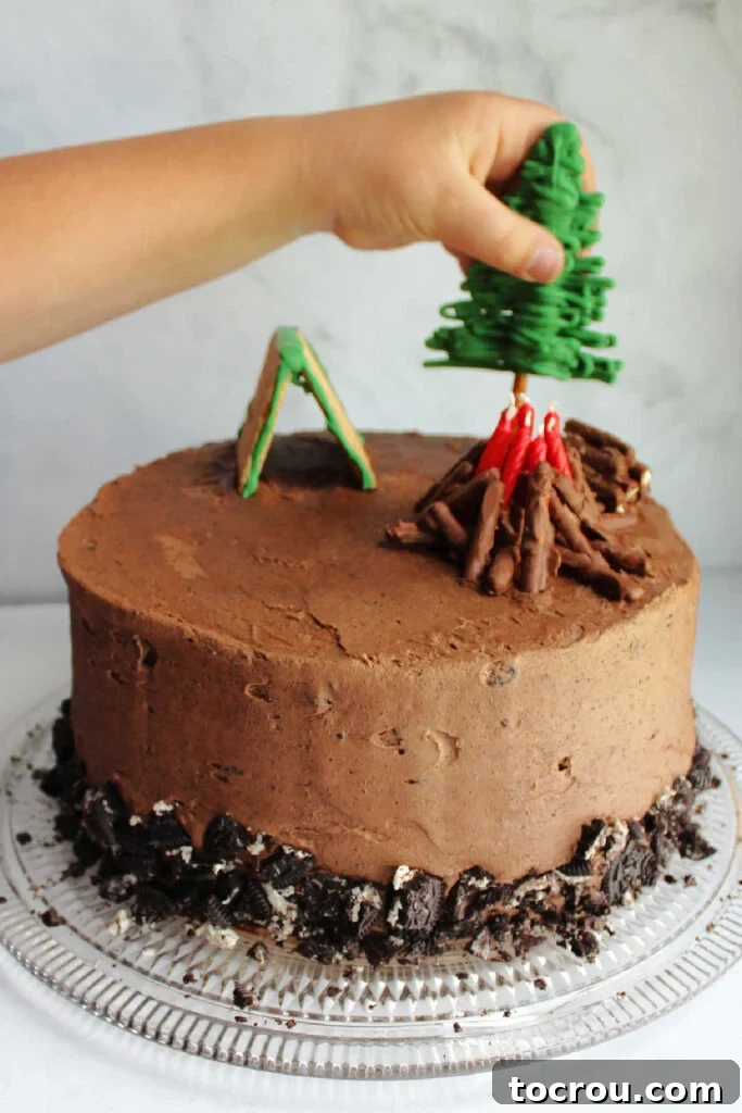 Adding Edible Decorations to Camping Cake Close-up of a child's hand carefully placing an edible pretzel tree into the frosted camping cake.