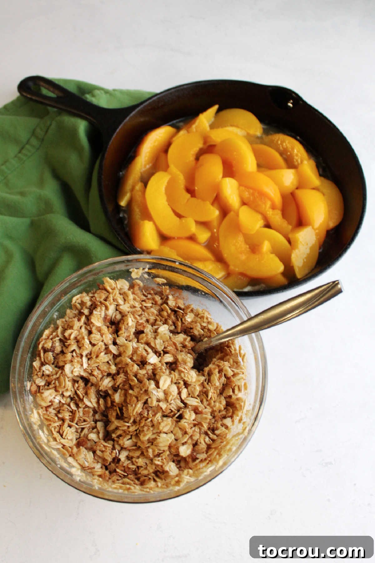 Cast iron pan filled with perfectly sliced peaches and a drizzle of maple syrup, with a bowl of crumbly crisp topping placed nearby.
