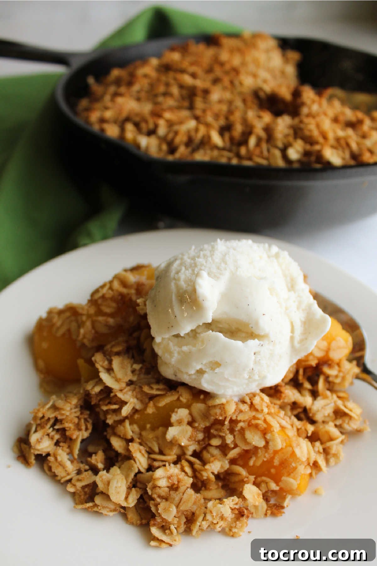 A serving of warm peach crisp with a scoop of melting vanilla ice cream in a white bowl, with the remaining crisp in the cast iron skillet in the soft-focused background.