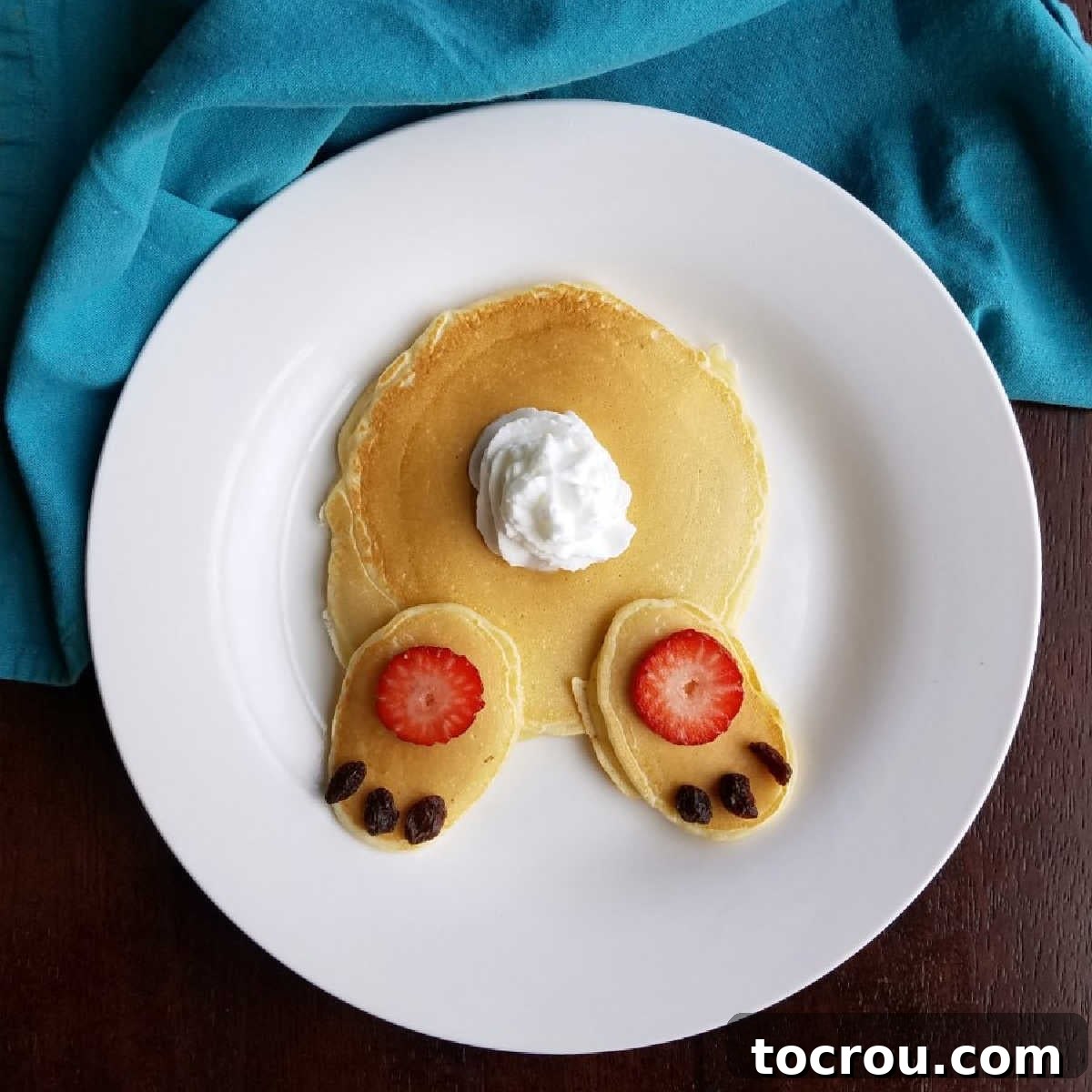 A breakfast plate featuring three homemade pancakes arranged to resemble a bunny's rear end, complete with whipped cream for a tail and sliced strawberries and raisins for feet.