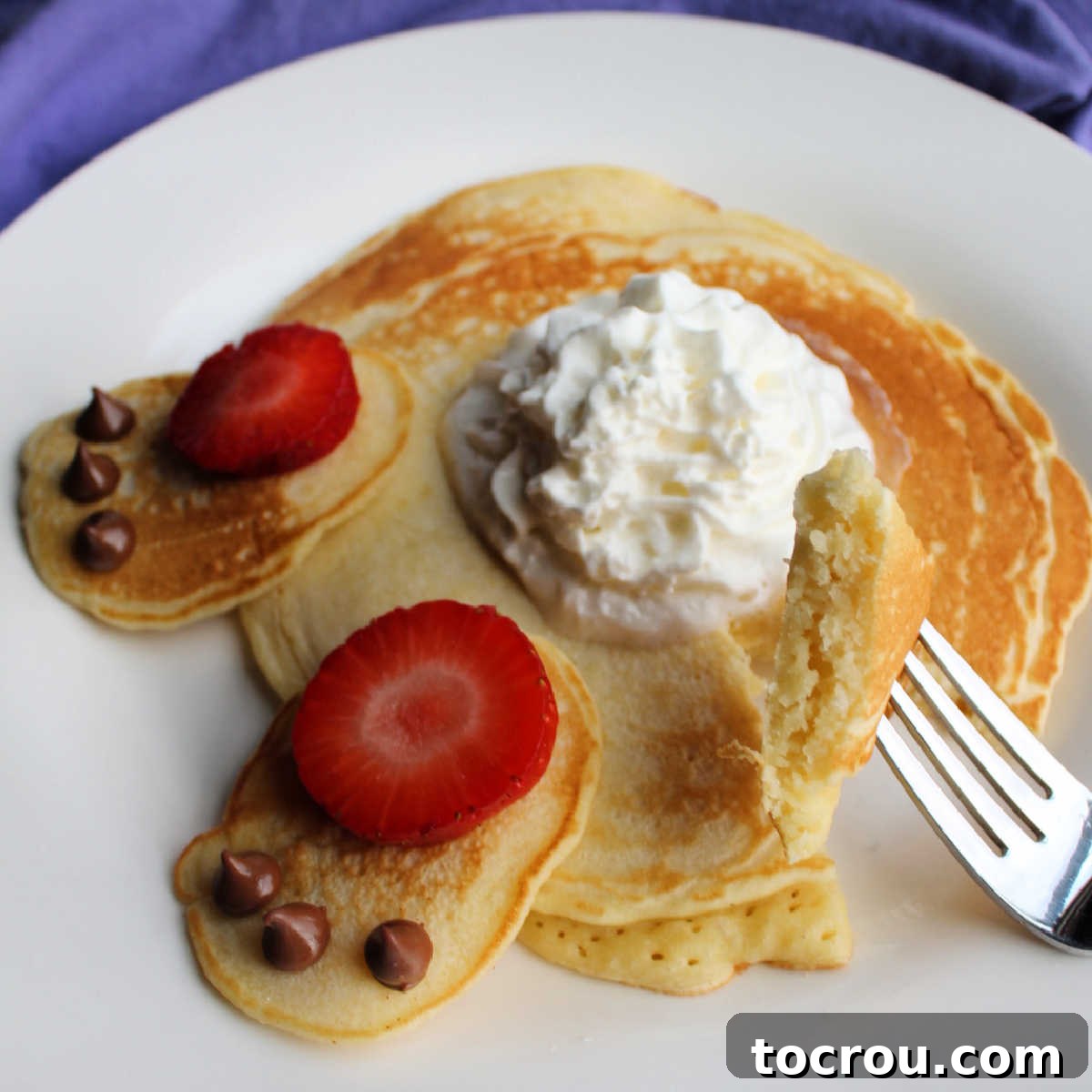 A detailed close-up reveals the soft, airy texture of homemade pancakes, with a fork holding a delicious bite. In the background, a whipped cream bunny tail and cheerful strawberry and chocolate chip bunny feet decorations add a festive touch.