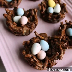 Treats shaped like birds nests made out of chocolate, peanut butter and crunchy chow mein noodles filled with pastel candy eggs on pink platter.