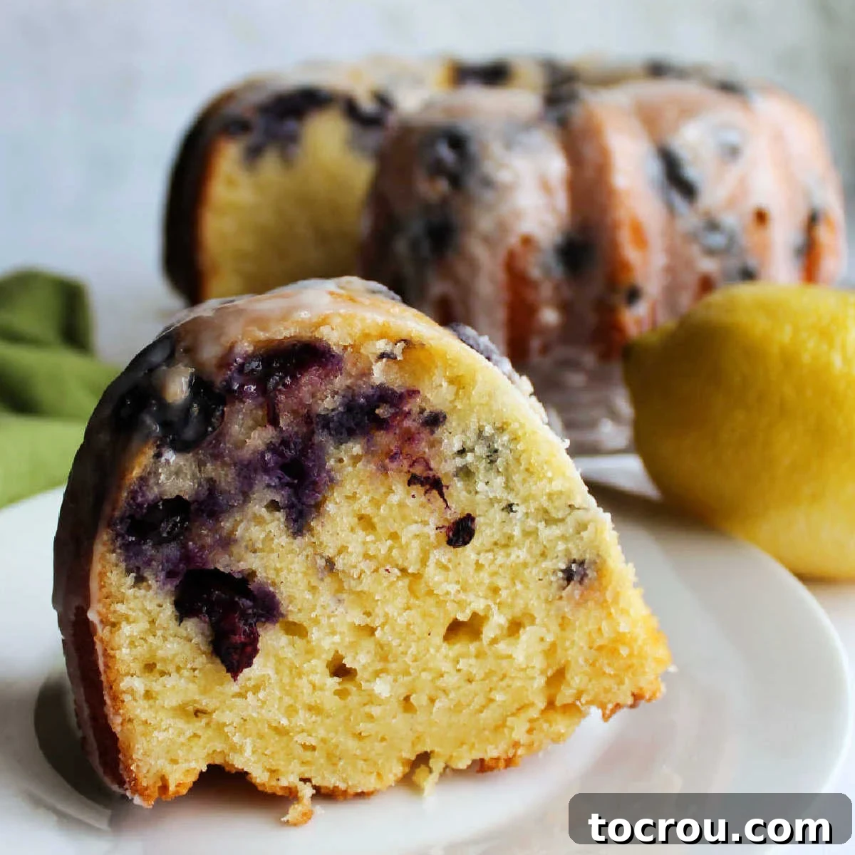 Lemon Blueberry Bliss Bundt 2 A dessert plate featuring a perfect slice of homemade lemon blueberry Bundt cake, with the rest of the cake, beautifully glazed with powdered sugar, in the background.