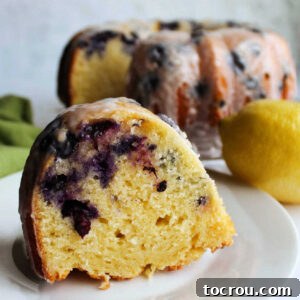 Lemon Blueberry Bliss Bundt 11 A dessert plate with a piece of homemade lemon blueberry Bundt cake on it with remaining cake drizzled with powdered sugar glaze in the background.