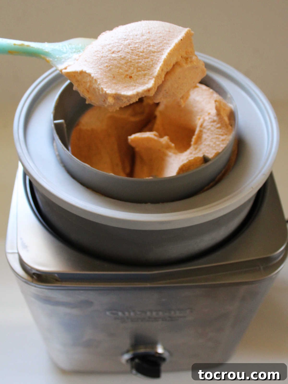 A spatula gently lifts a generous portion of freshly churned pumpkin ice cream from the ice cream machine, showcasing its creamy texture and inviting color before it's moved to storage.