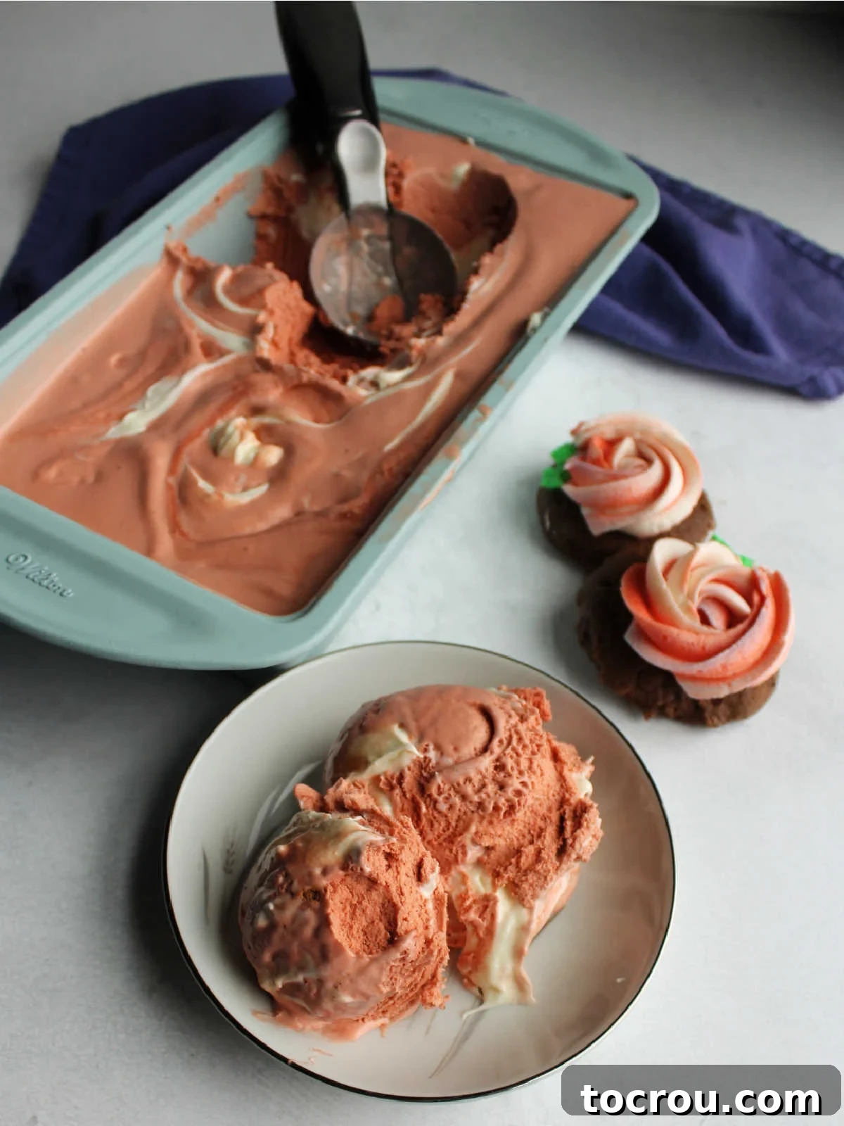 A beautiful bowl filled with scoops of pink velvet ice cream, with a loaf pan containing the remaining ice cream and frosted chocolate cookies visible in the softly blurred background.