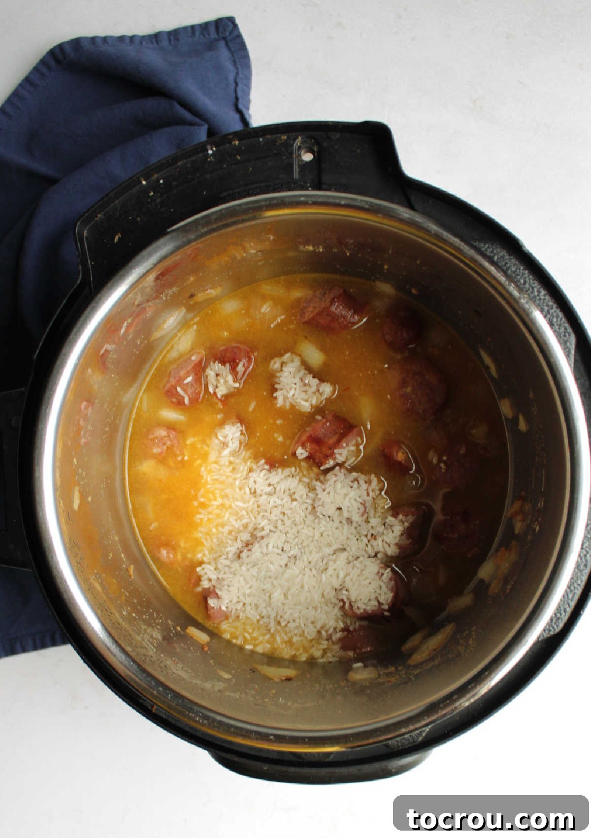 Inside the Instant Pot, uncooked long-grain rice and clear chicken broth have been added to the browned kielbasa and sautéed onions, forming the next layer of the one-pot dish, ready for pressure cooking.