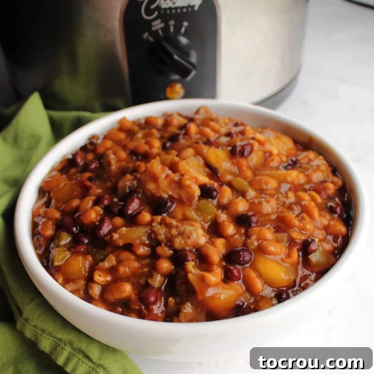 Ultimate Slow-Cooked Baked Beans 2 A serving bowl filled to the brim with loaded baked beans, rich with sauce, bacon, and sausage, positioned invitingly in front of a slow cooker.
